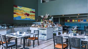 Un restaurante moderno con una barra central, varias mesas para comer y sillas. Hay un cuadro abstracto y colorido en la pared, y una variedad de plantas y flores en la barra.