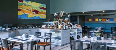 Un restaurante moderno con una barra central, varias mesas para comer y sillas. Hay un cuadro abstracto y colorido en la pared, y una variedad de plantas y flores en la barra.