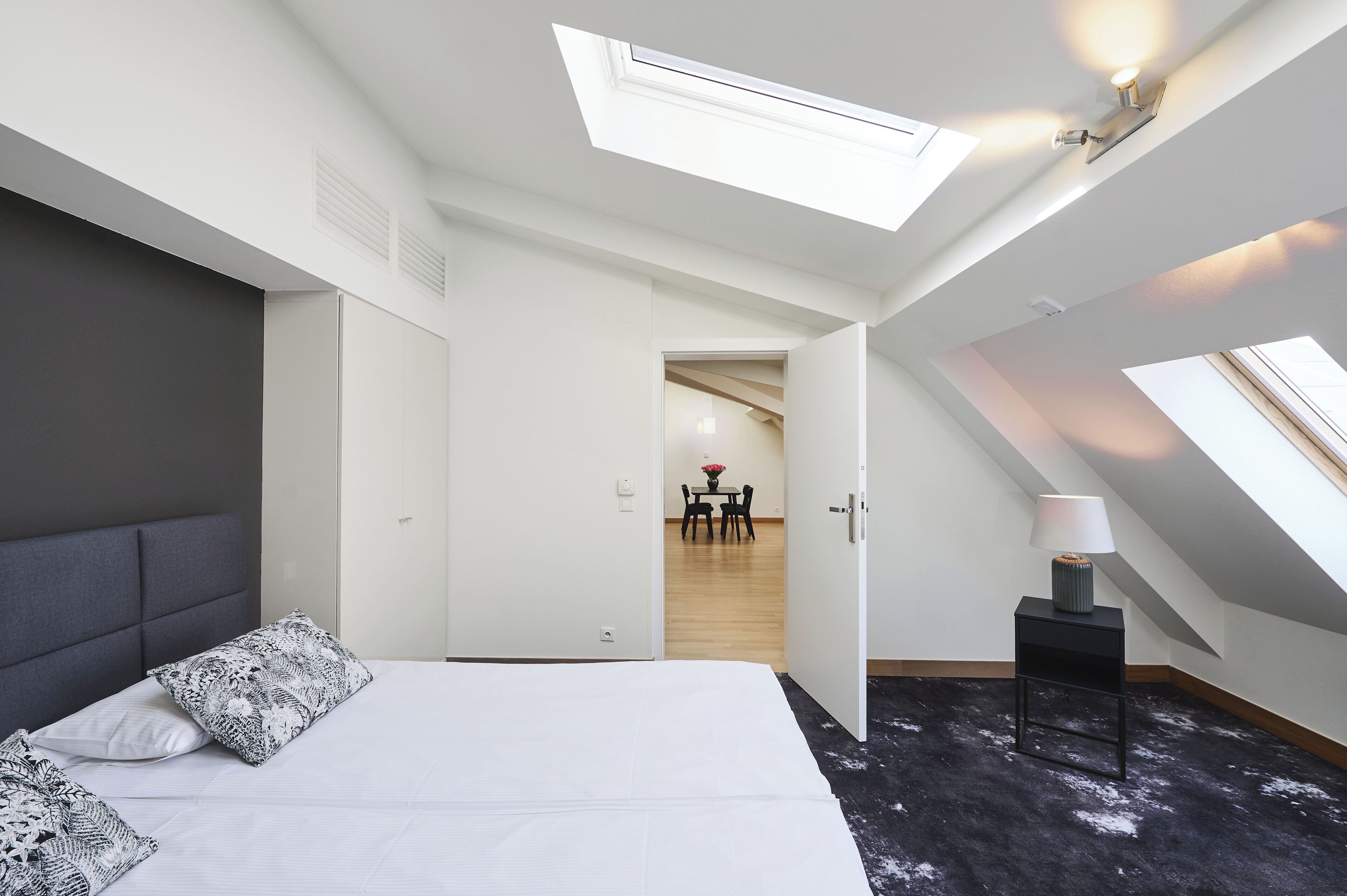 A bedroom with a bed, a bedside table with a lamp, a skylight, and a view of a dining area.