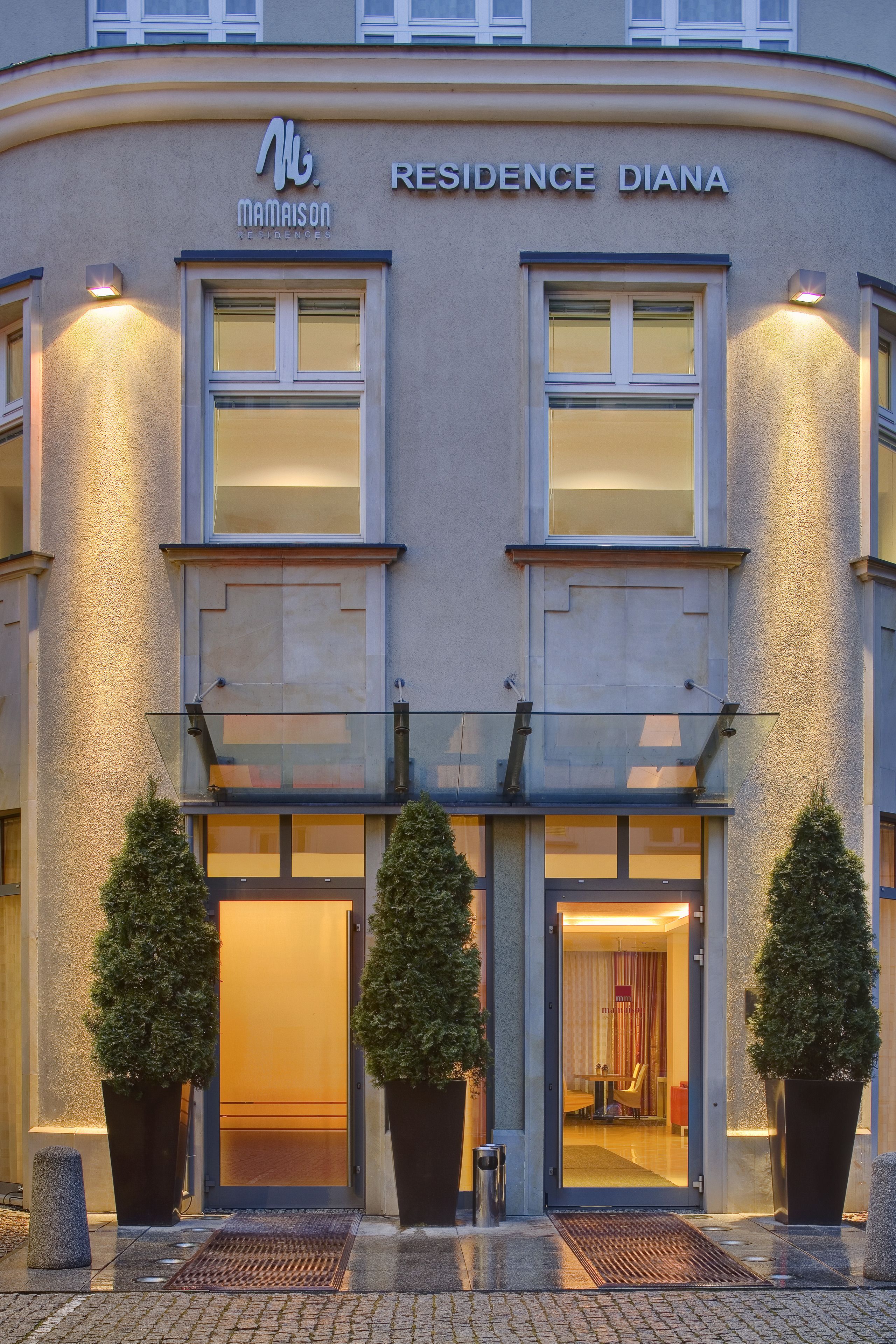 A modern building with a sign reading "Residence Diana." It features a glass entrance, potted plants, and a well-lit facade.