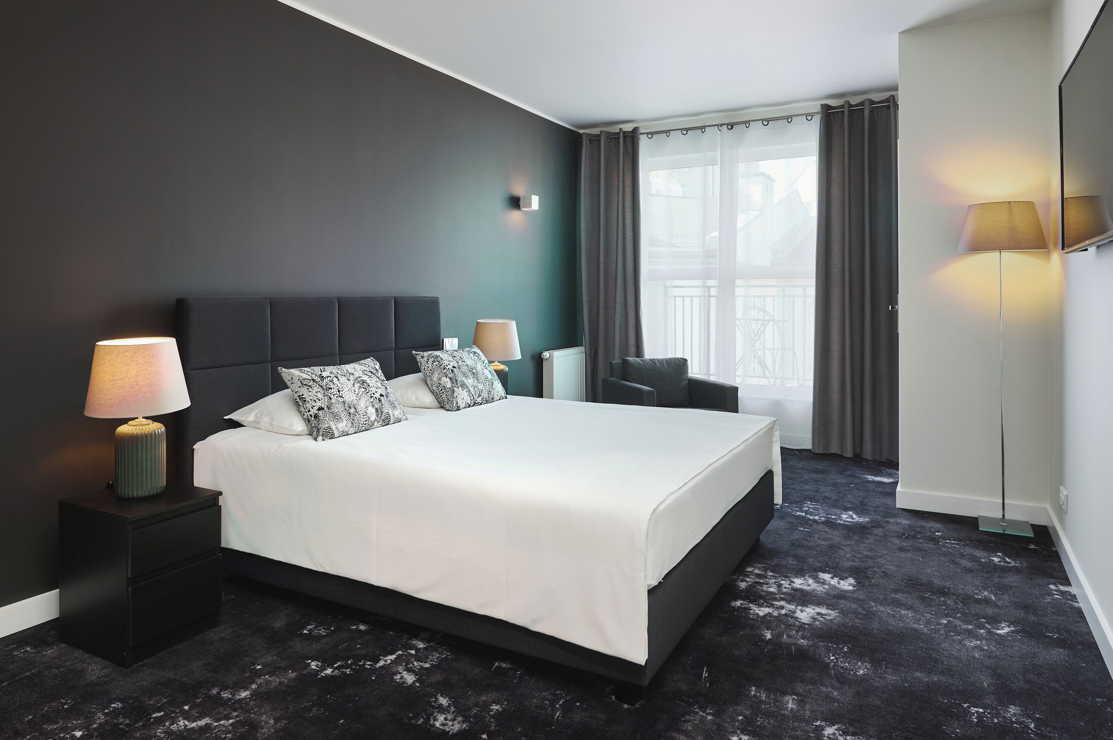 A modern hotel room with a large bed, bedside lamps, a dark headboard, and a view of the outside through large windows.