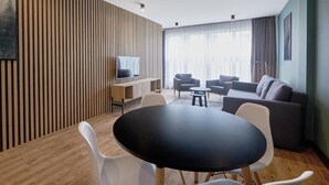 A modern living room with a wooden slatted wall, a flat-screen TV, a round black table with white chairs, a grey sofa, and a large window with sheer curtains. - Mamaison Residence Diana (Warsaw)