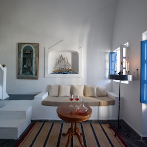 A modern living room with a beige sofa, a small wooden table with two wine glasses, a ship model, and a painting on the wall. - Heliotopos Hotel (Santorini)