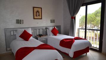 Habitación de hotel con dos camas, cada una con ropa blanca y detalles rojos, una mesita de noche con teléfono, un cuadro en la pared y un balcón con vista a árboles.