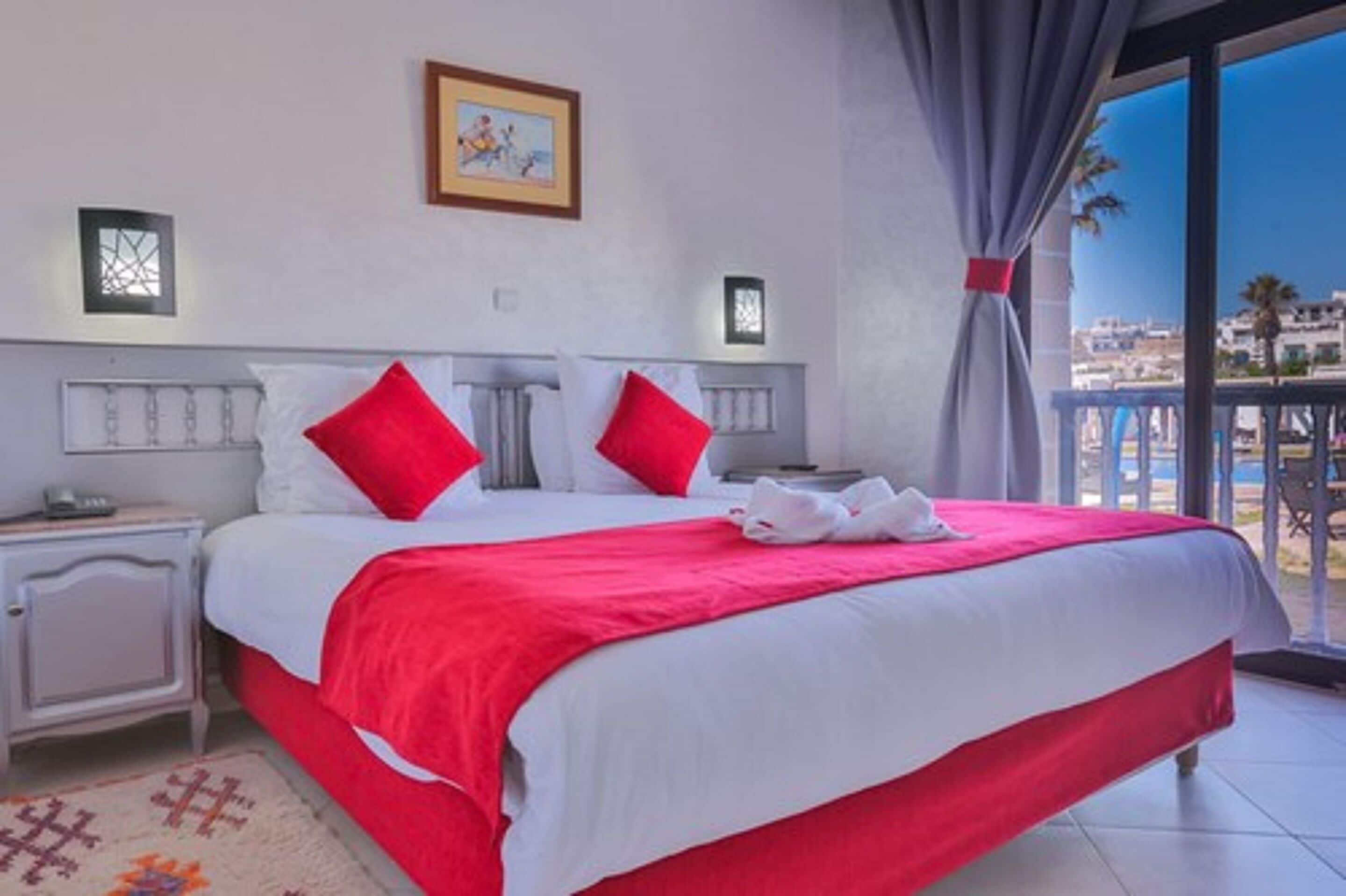 Een hotelkamer met een groot bed, witte nachtkastjes, rood-witte beddengoed, een inlijsting foto aan de muur, en een balkon met uitzicht op de stad.