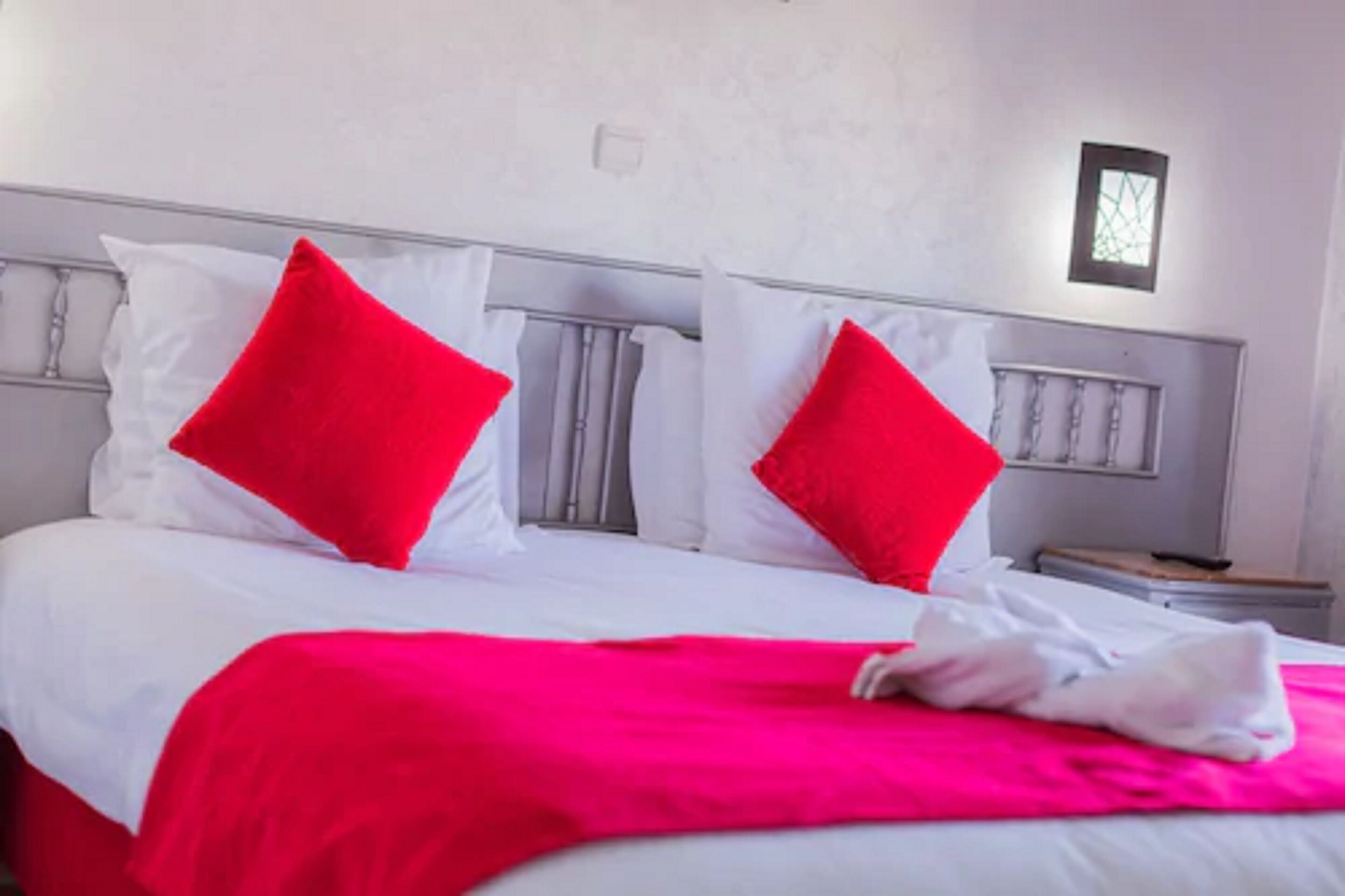 Ein Bett mit weißer Bettwäsche, zwei roten Kissen, einer hellrosa Decke und einem weißen Handtuch.