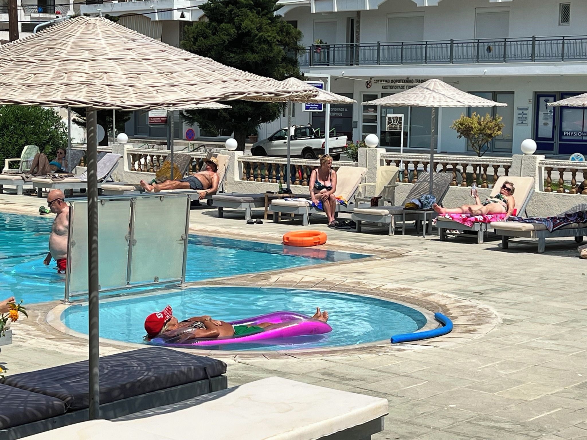 Ein Poolbereich mit Liegestühlen und Sonnenschirmen, ein Schwimmbecken und ein Gebäude mit Beschilderung im Hintergrund.