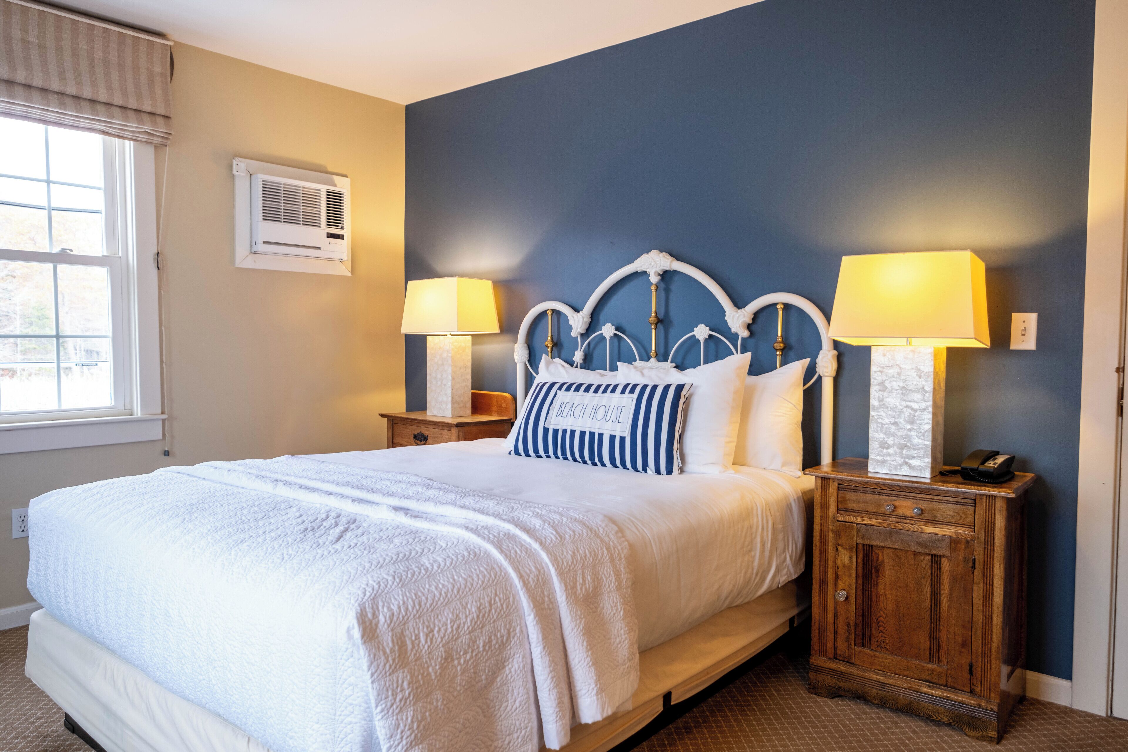 Una cama bien hecha con cubrecama y almohadas blancas, dos lámparas de noche, una mesita de madera y una ventana con persianas.