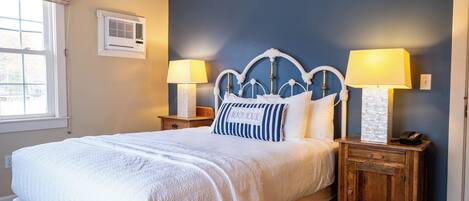 Una cama bien hecha con cubrecama y almohadas blancas, dos lámparas de noche, una mesita de madera y una ventana con persianas.