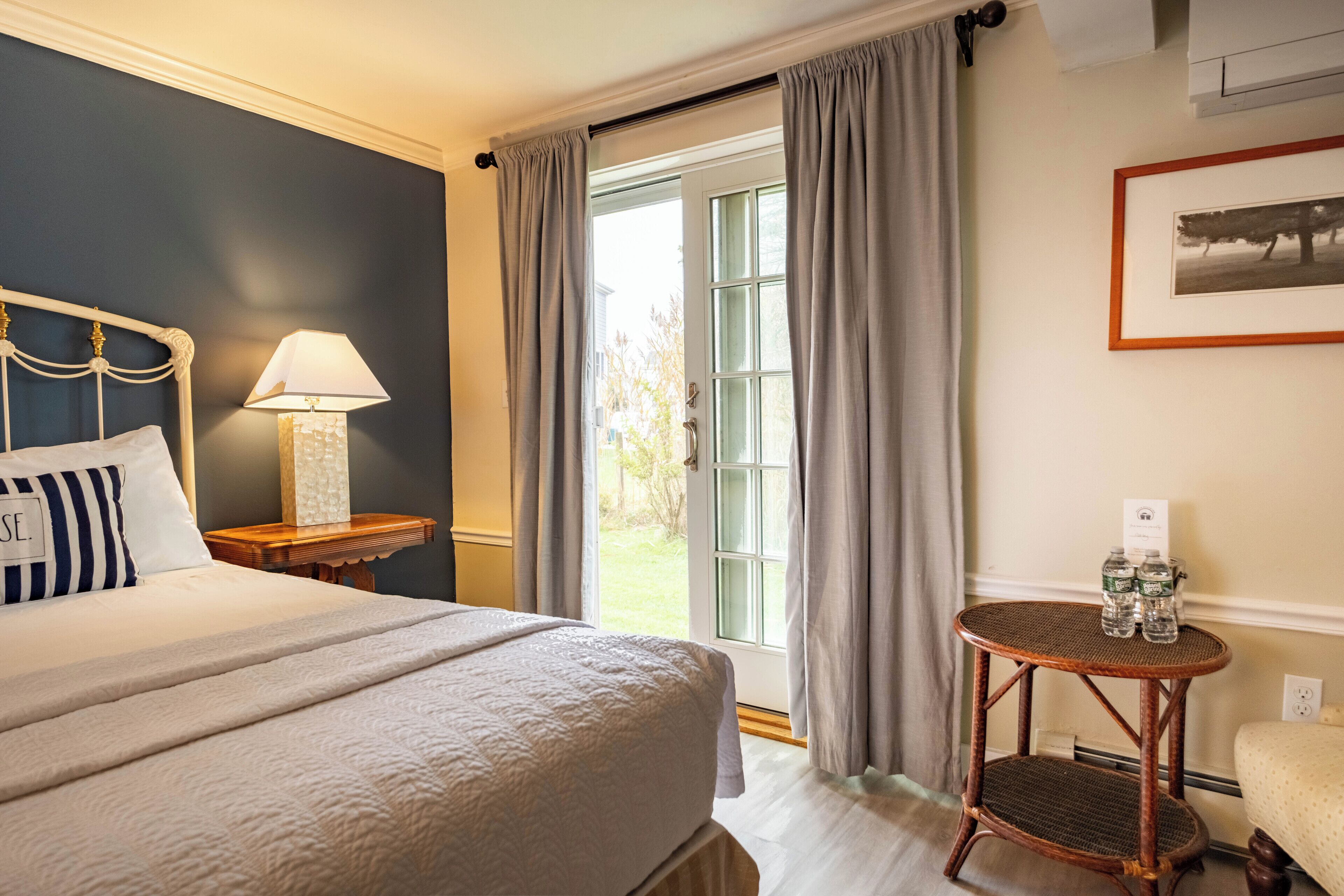 A hotel room with a bed, a bedside table with a lamp, a small round table with bottles, and a window with curtains.