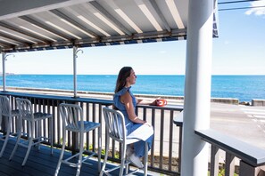 Une personne assise sur un balcon, avec vue sur la mer, des chaises blanches et une tasse rouge.
