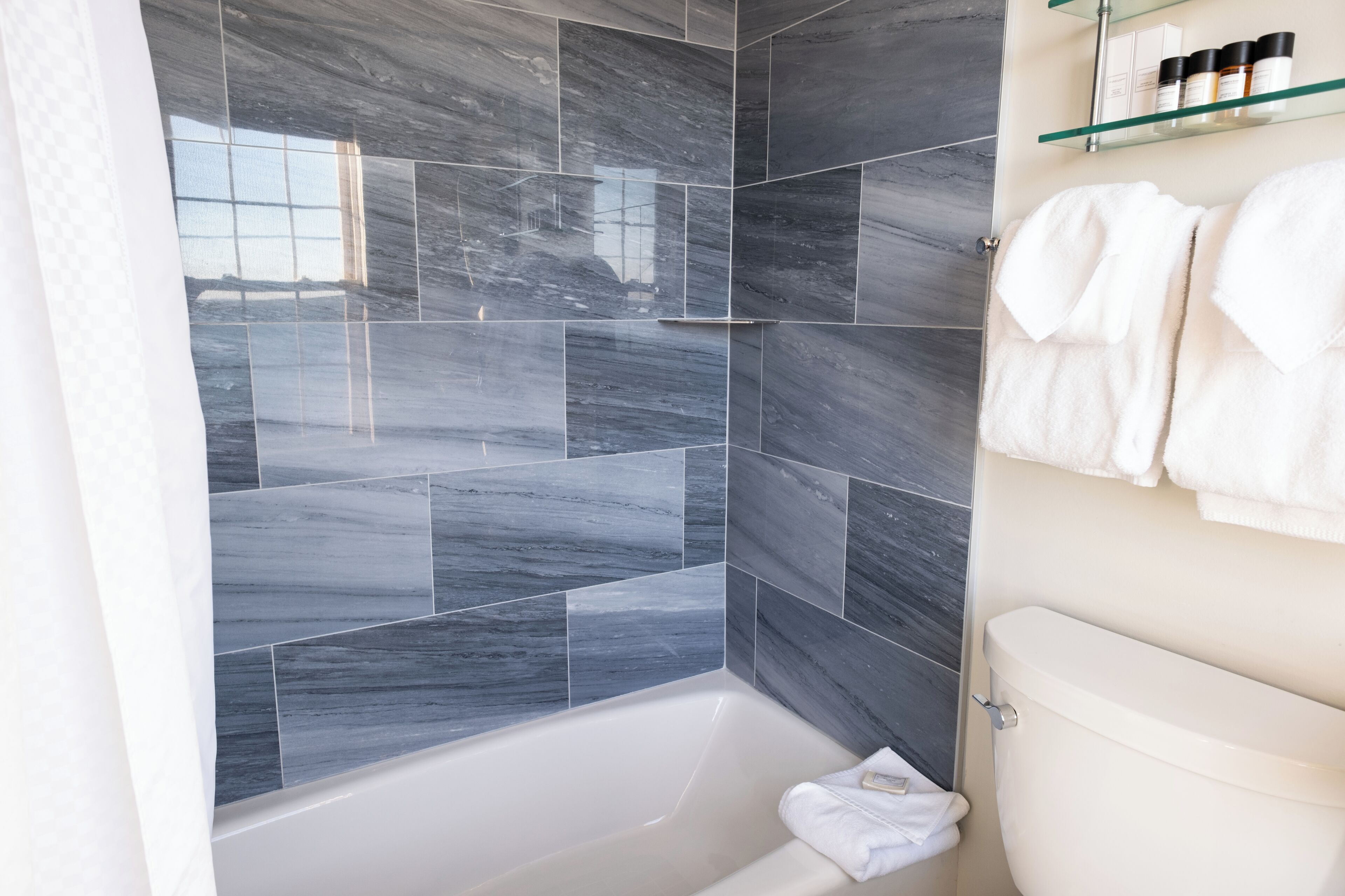 A bathroom with a bathtub, dark tiled walls, and a glass shelf with toiletries.