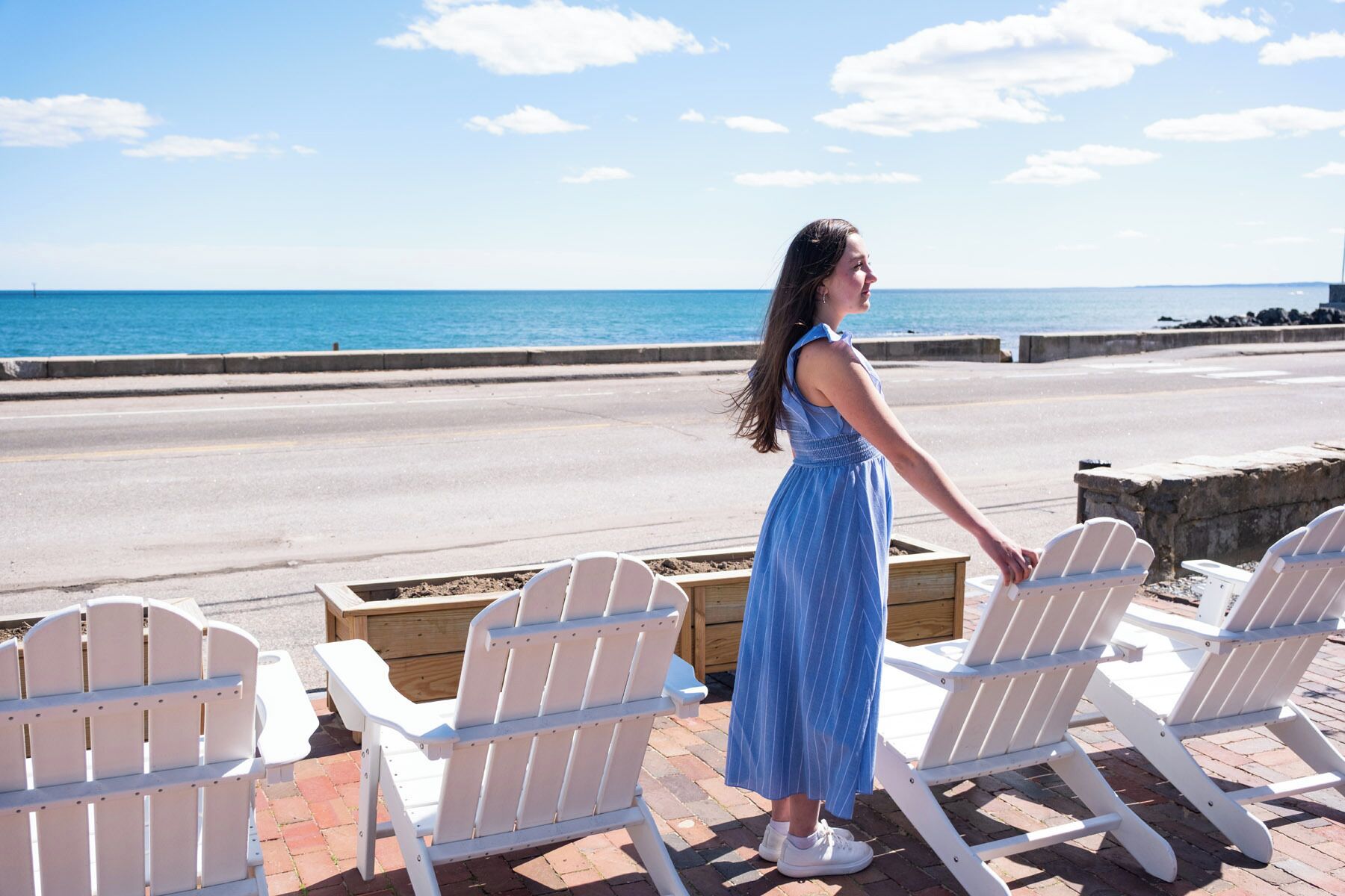 A woman standing by a seaside with white Adirondack chairs and a wooden planter.