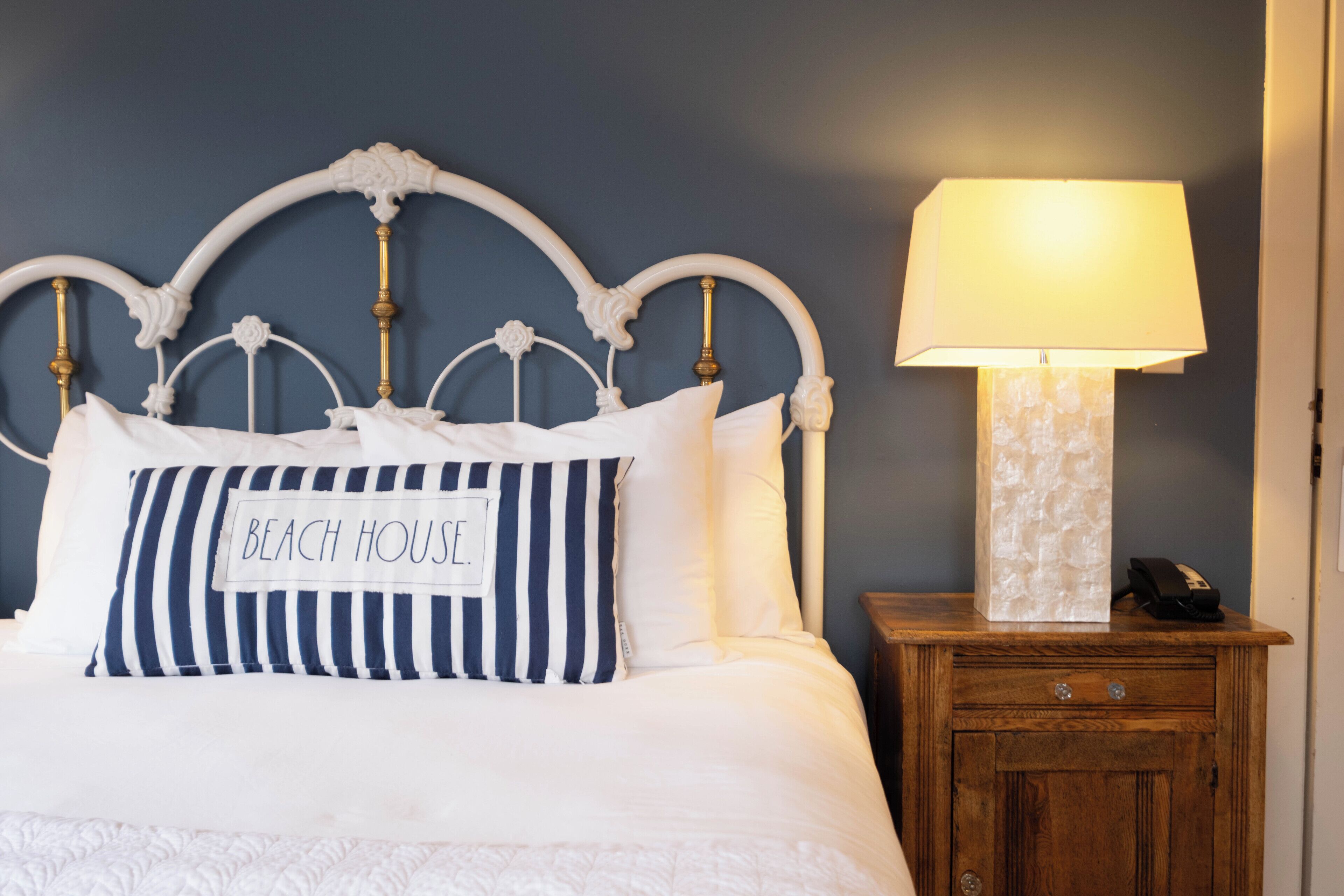 A neatly made bed with a pillow labeled 'BEACH HOUSE' and a bedside table with a lamp.