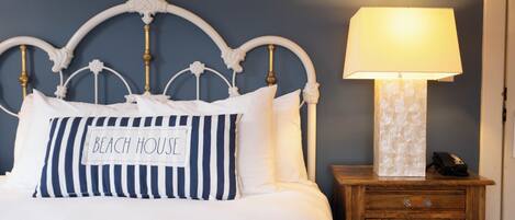 A neatly made bed with a pillow labeled 'BEACH HOUSE' and a bedside table with a lamp.