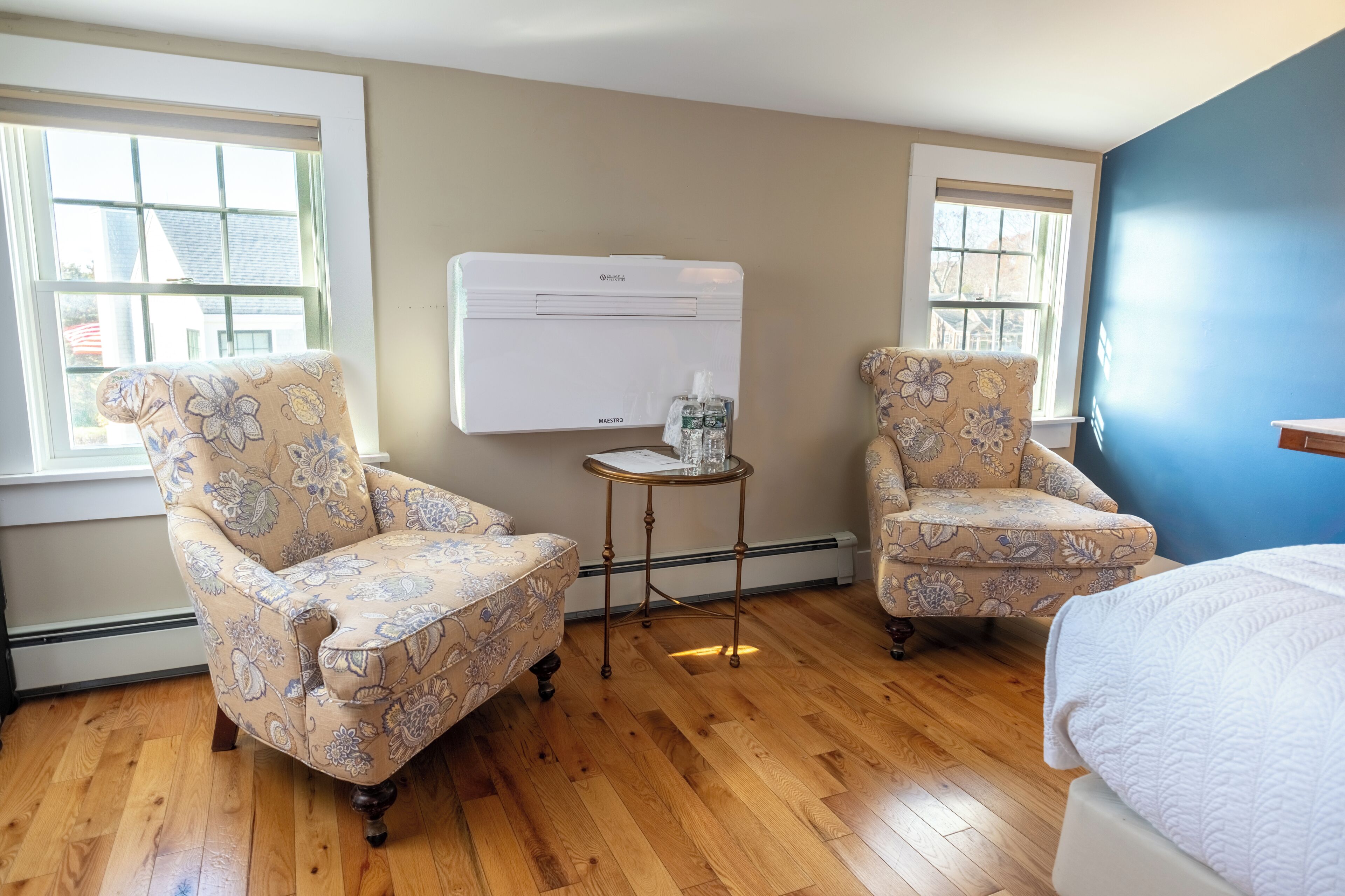 Una habitación con dos sillones de estampado floral, una mesita redonda con una botella y un vaso, y una cama con colcha blanca.