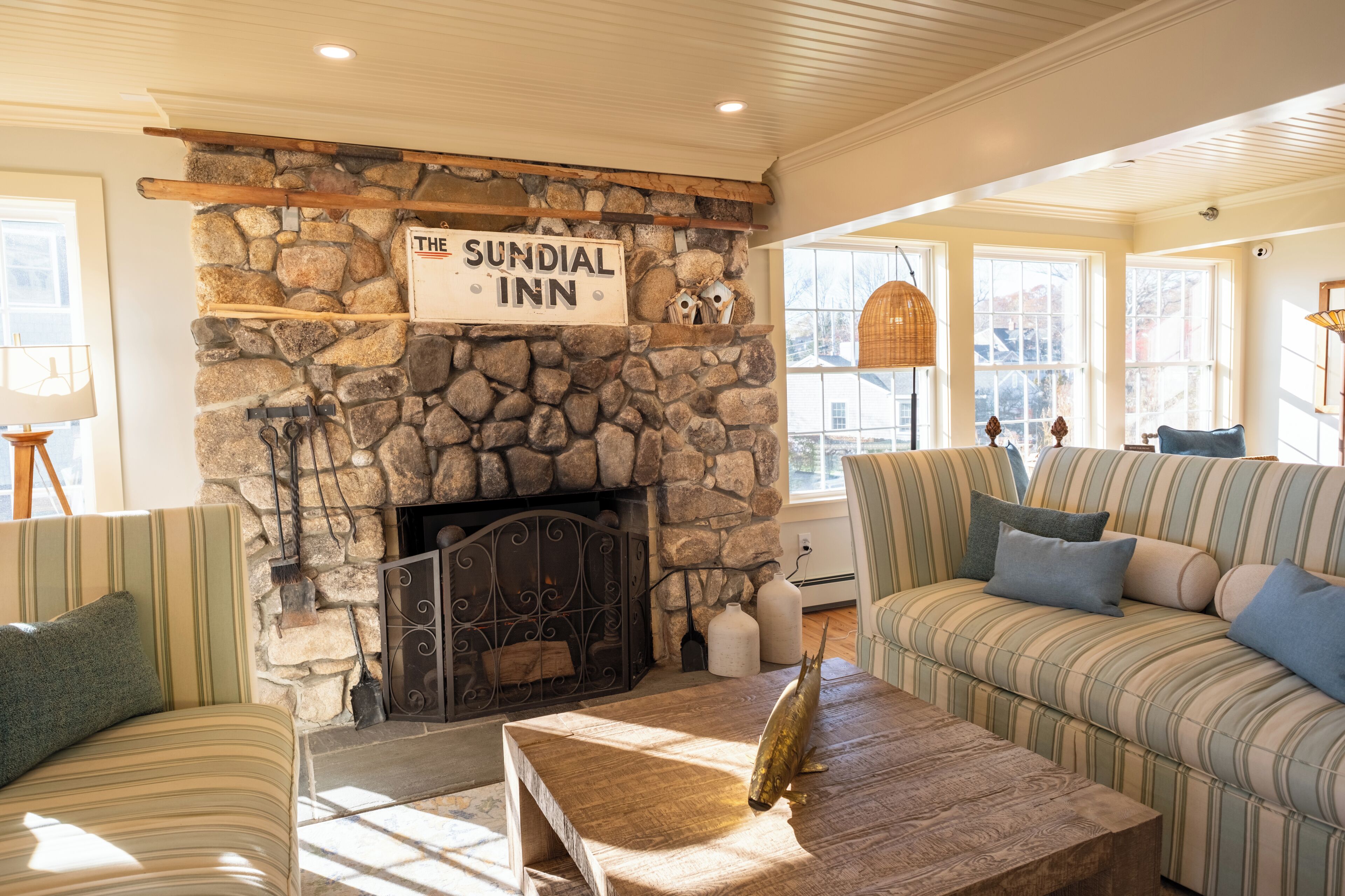 A cozy room with a stone fireplace, a sign reading "The Sundial Inn," striped sofas, and a wooden coffee table.