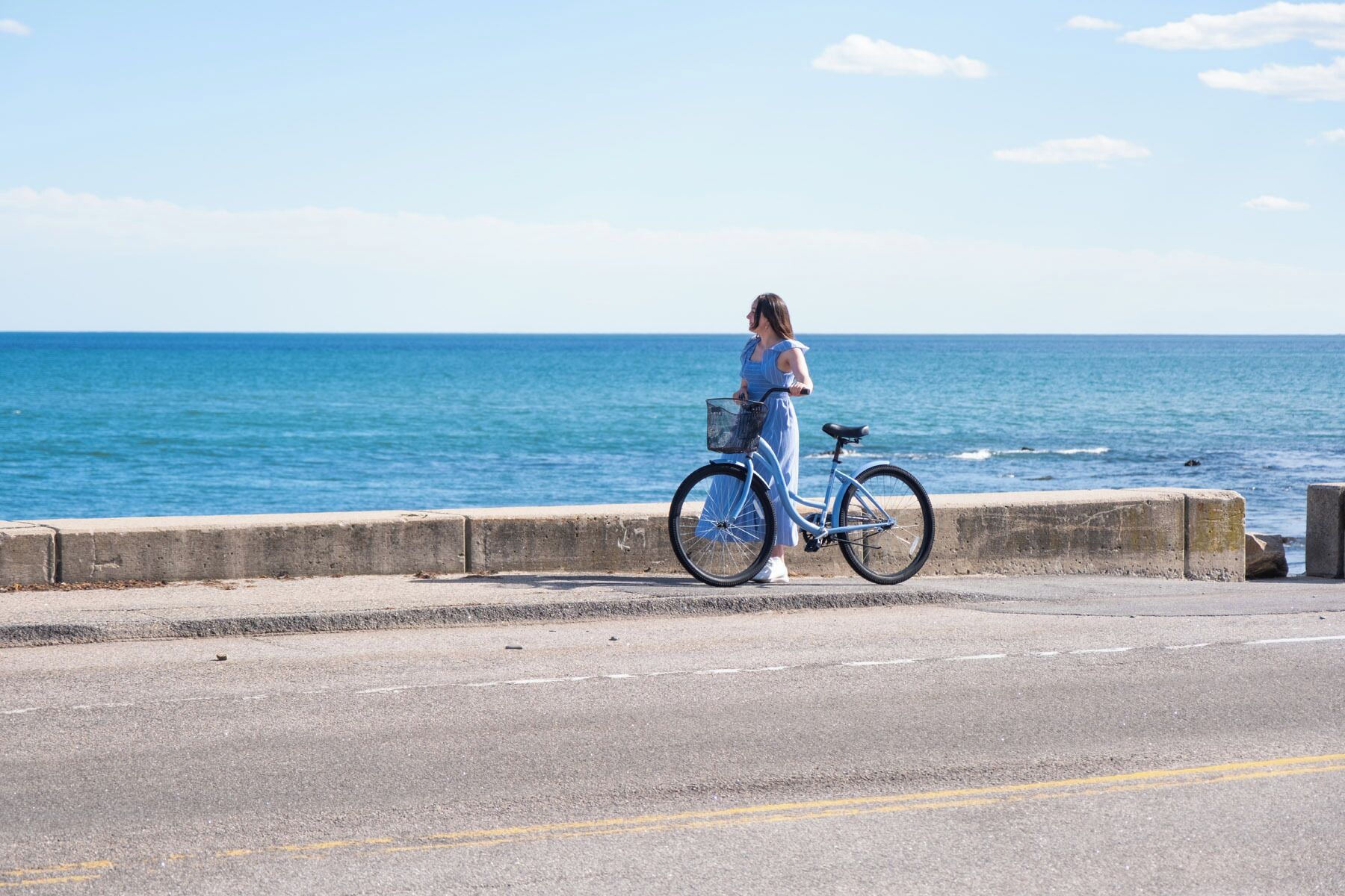 A coastal road with a concrete barrier, a person standing next to a bicycle, and the ocean in the background.