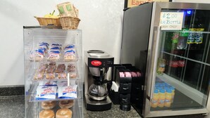 Una estación de bebidas autoservicio con un cartel de precios, una máquina de café y un refrigerador con bocadillos y bebidas.