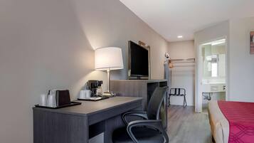 A hotel room with a desk, a chair, a bed, a TV, a lamp, and a small table with a coffee maker and cups.
