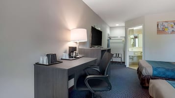 A hotel room with a desk, chair, bed, TV, and a small bathroom.