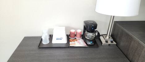 Mesita de noche en una habitación de hotel con cafetera, lámpara y bandeja de artículos de aseo.