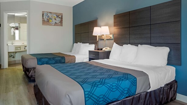 Habitación de hotel con dos camas, una mesita de noche con lámpara, baño visible a través de una puerta entreabierta y una obra de arte colgada en la pared.