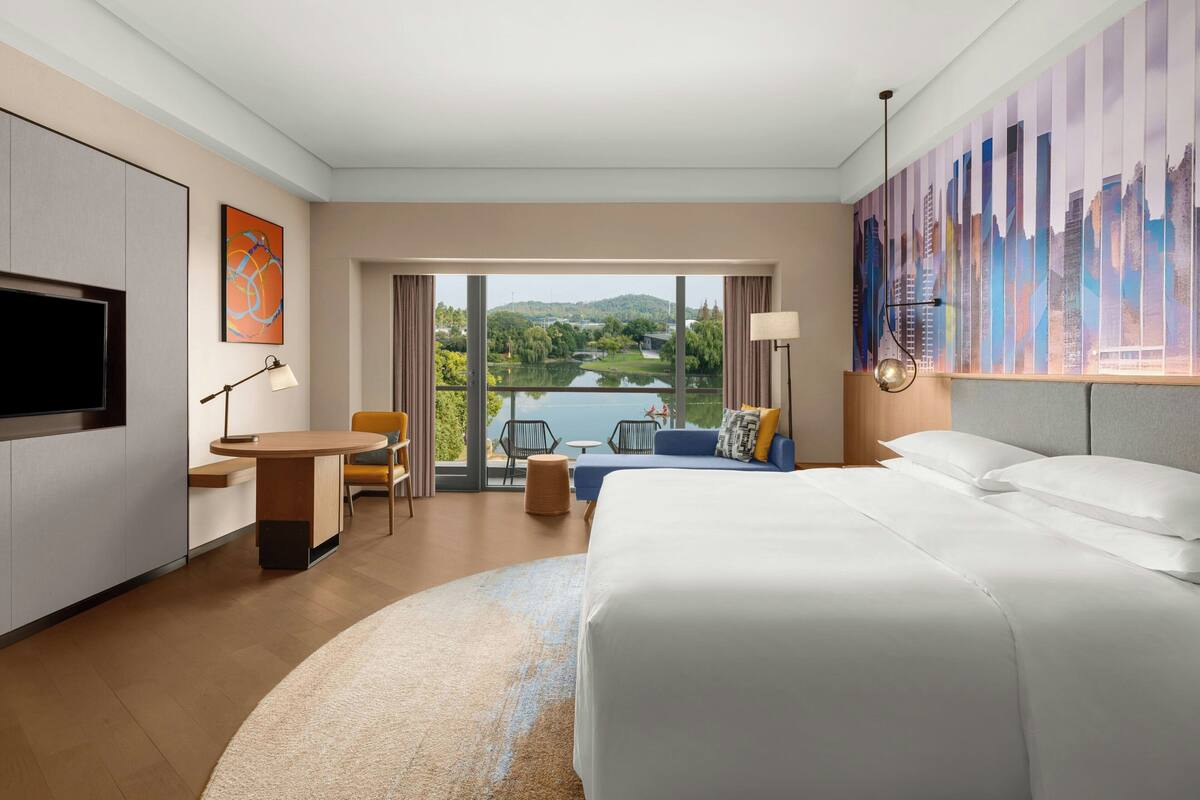 A modern hotel room with a large bed, a desk, a chair, and a view of a lake and cityscape.
