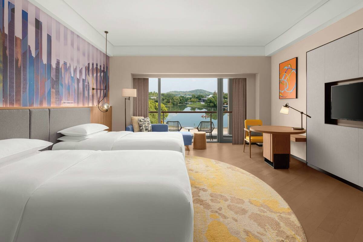 A hotel room with a large bed, a small desk, a chair, and a TV. There is a view of a lake and hills outside the window.