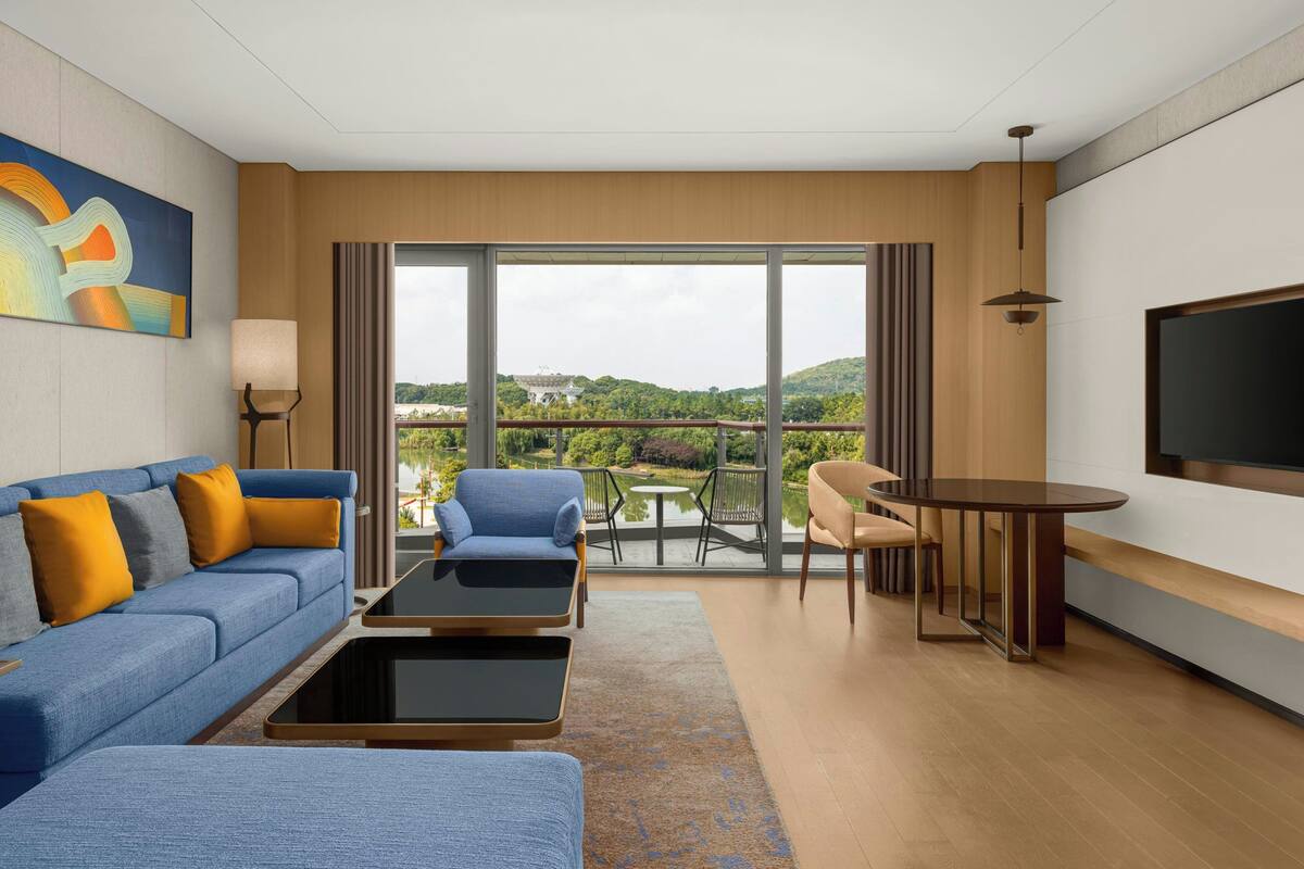 A modern hotel room with a sofa, two armchairs, a small table, a TV, and a balcony with a view of greenery and a building.