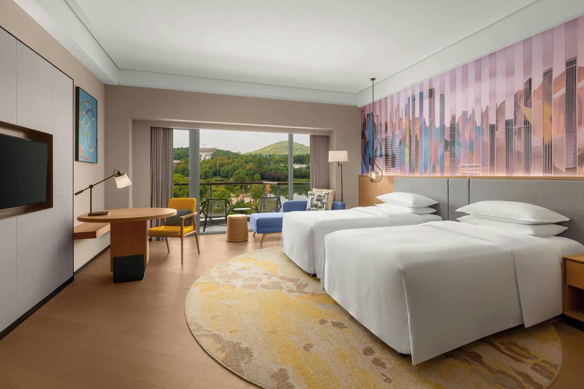 A hotel room with two beds, a TV, a dining area, and a balcony with a cityscape view.