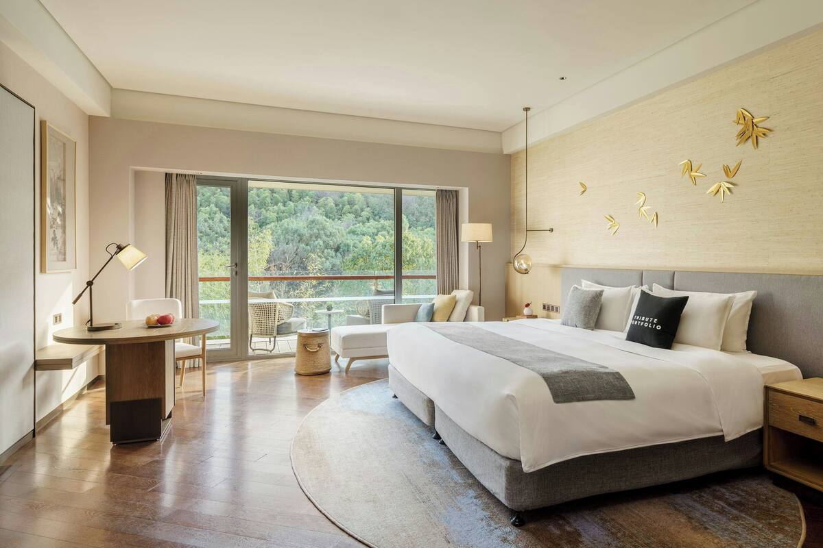 A modern hotel room with a large bed, a desk, a chair, and a view of the outdoors.