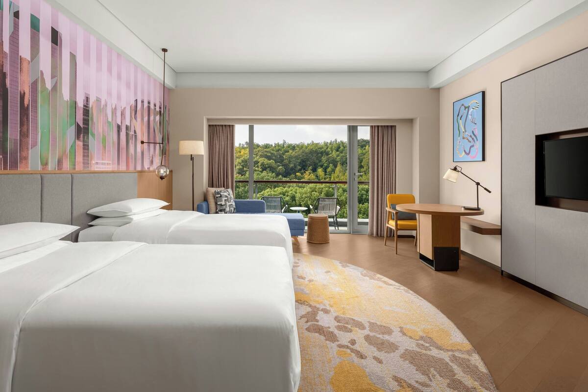 A hotel room with two beds, a desk, a chair, a TV, and a balcony with a view of trees.