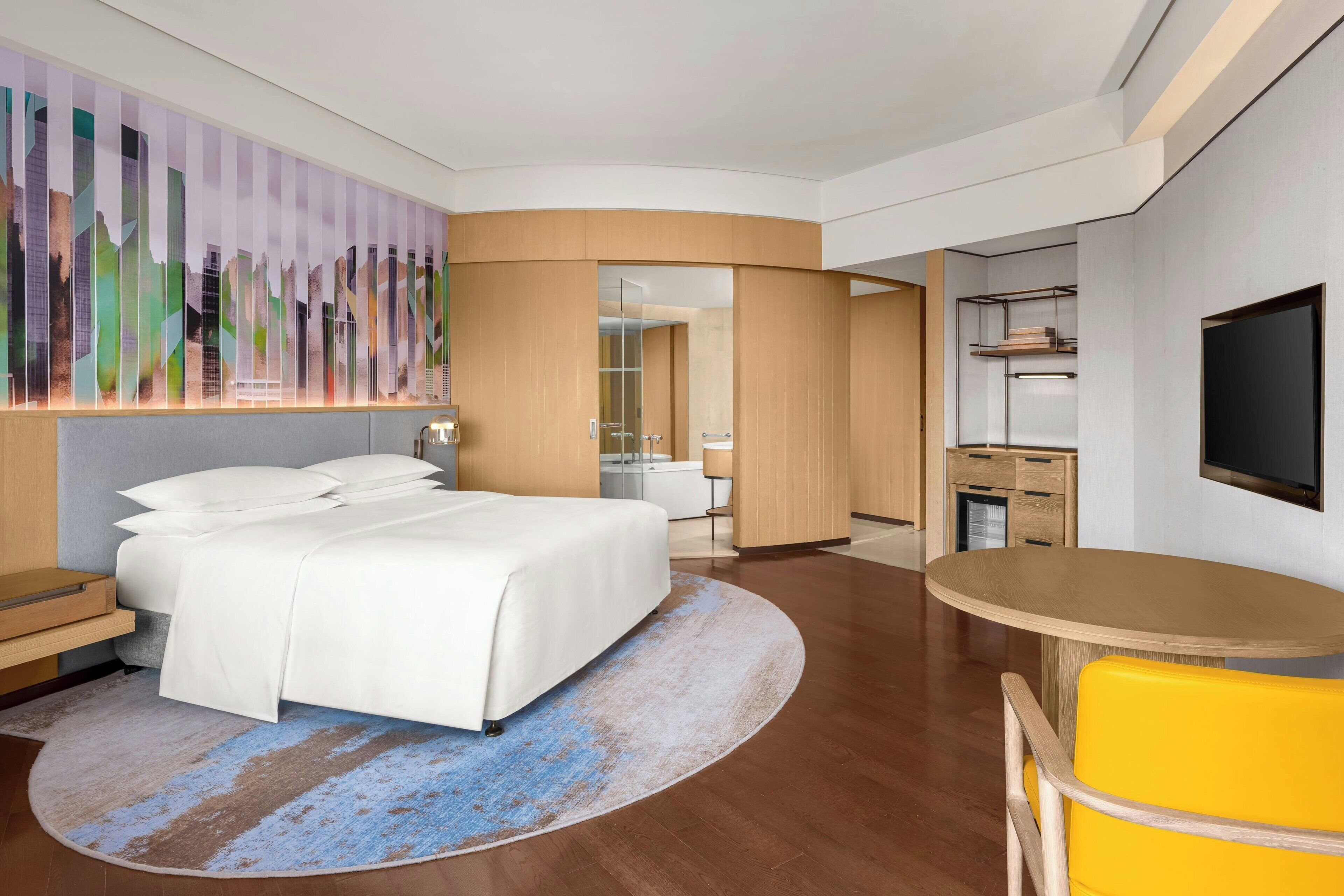 Habitación de hotel moderna con una cama grande, un escritorio, una silla, un televisor y un baño visible a través de una puerta abierta.