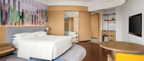 Habitación de hotel moderna con una cama grande, un escritorio, una silla, un televisor y un baño visible a través de una puerta abierta.