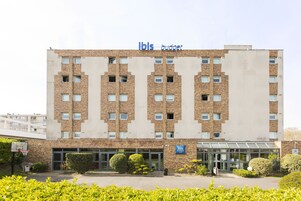 A multi-story building with a sign reading "Ibis Budget." It features a brick facade, a glass entrance, and a basketball court in front.