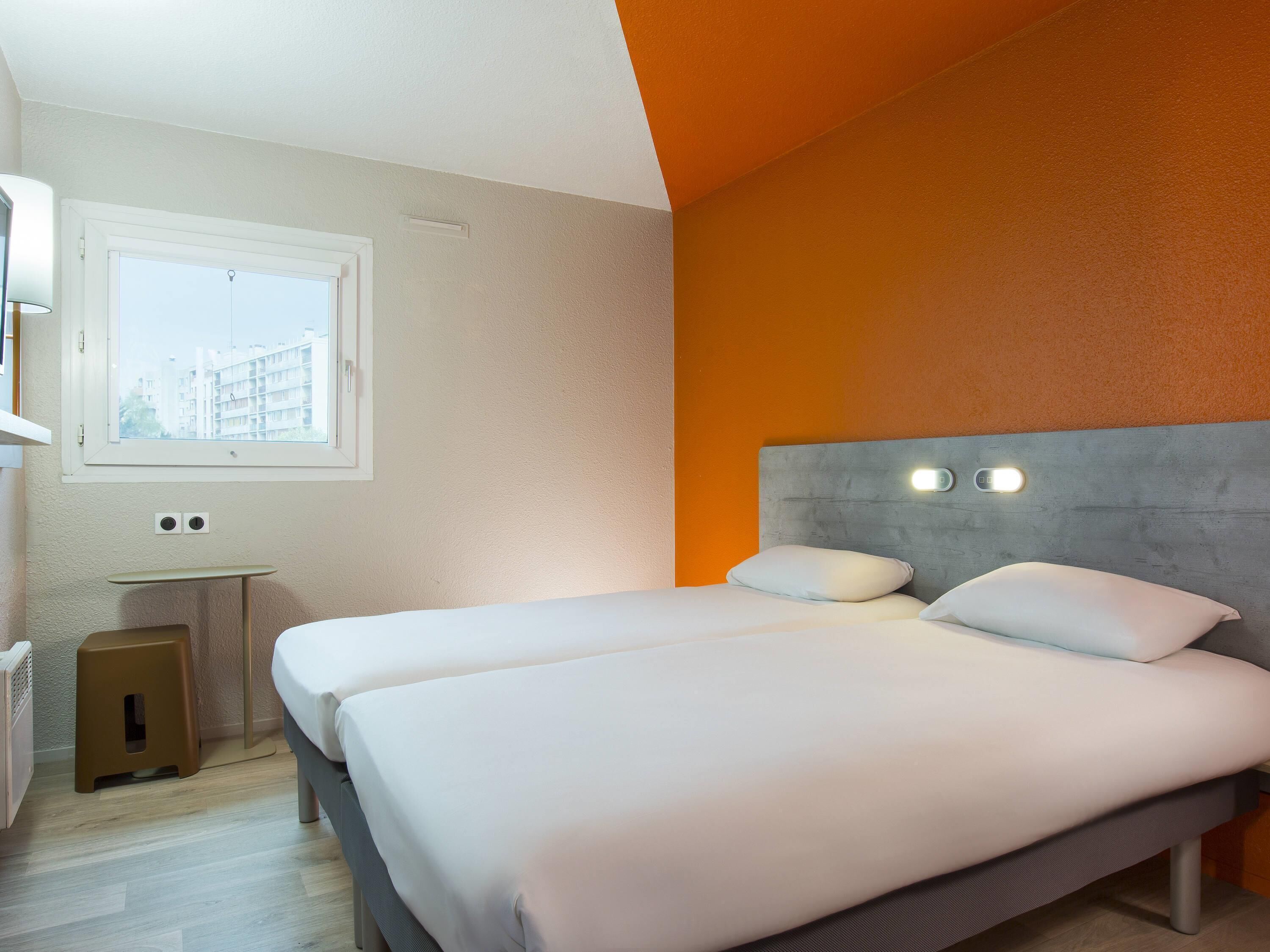 A hotel room with two beds, a window, a bedside table, and a small stool.