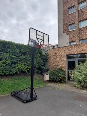 A basketball hoop with the brand name "TARMAK" in front of a brick building with a green hedge and a yellow flowering plant nearby. - Ibis Budget Fresnes (Fresnes)