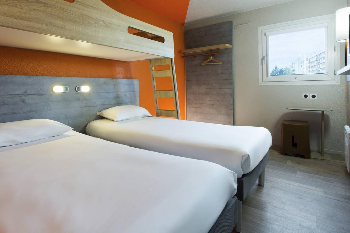 A hotel room with two beds, a window, a small table, and a wall-mounted shelf.
