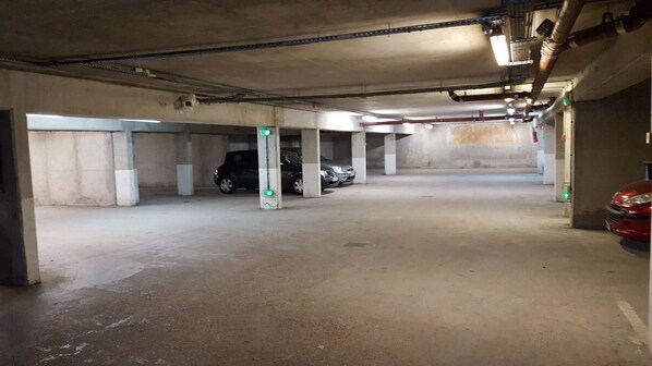Un parking avec des piliers en béton, quelques voitures garées et des tuyaux apparents au plafond.