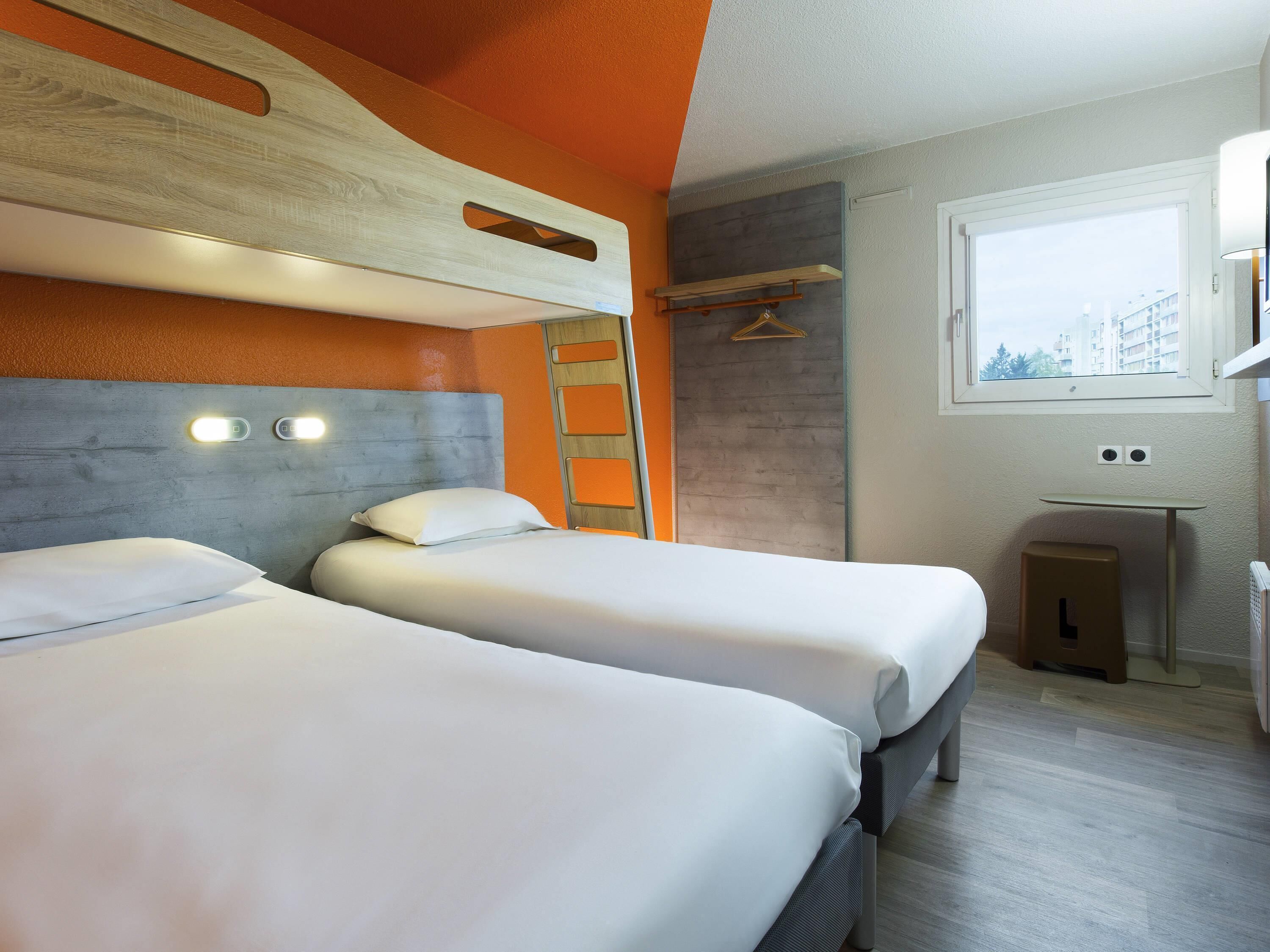 A hotel room with two beds, a window, a small table, and a wall-mounted shelf.