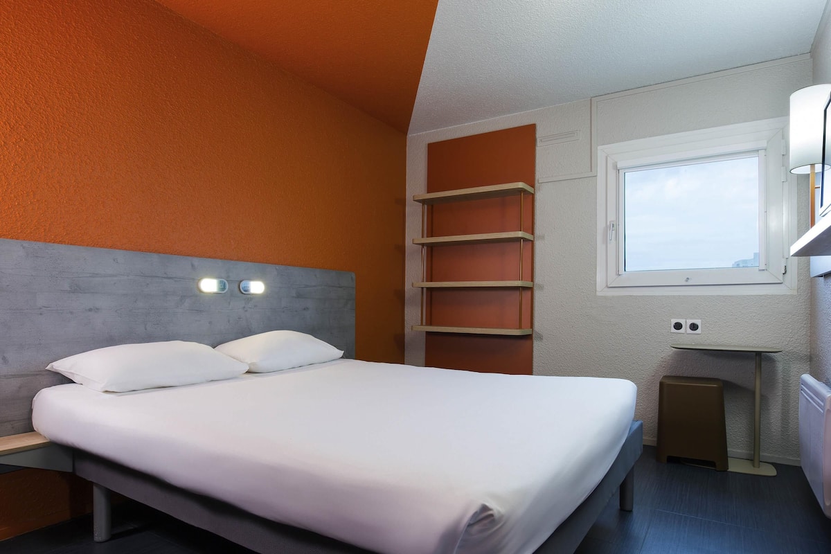 A modern hotel room with a bed, a nightstand, a window, and built-in shelves.