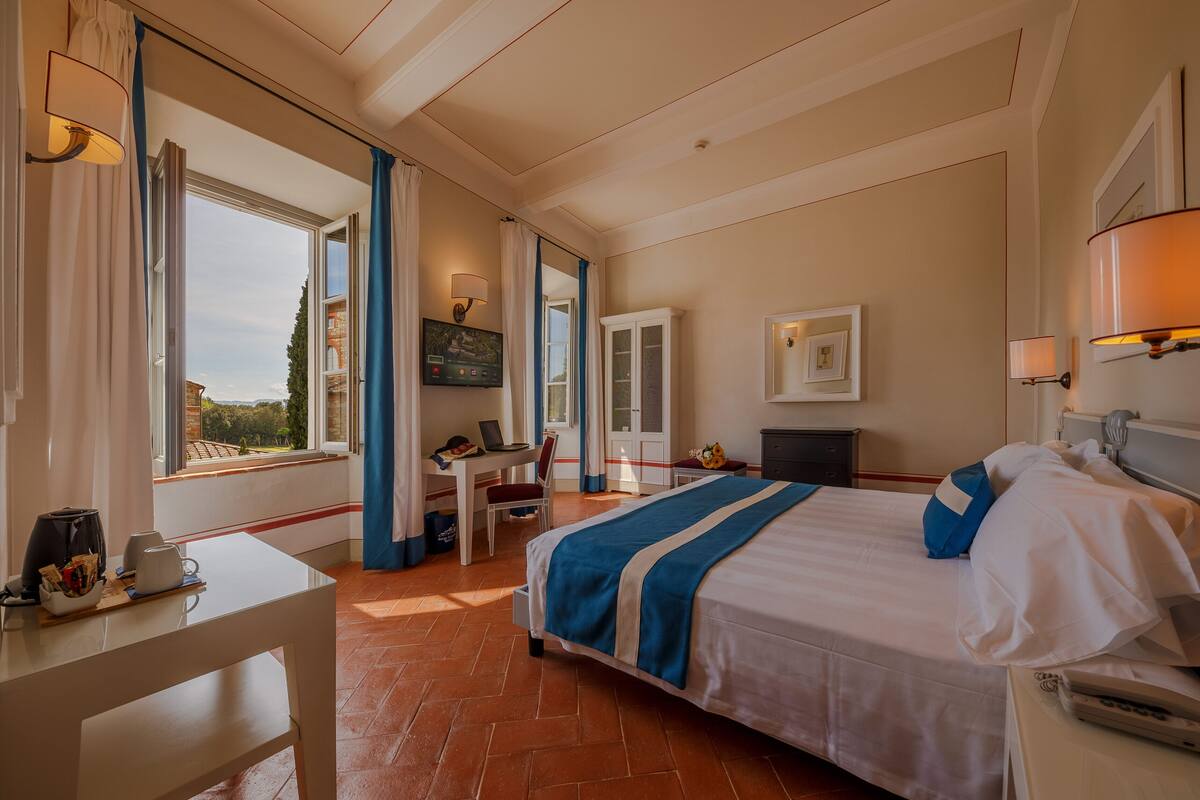 Een hotelkamer met een bed, bureau en stoel, een raam met gordijnen, en uitzicht op buiten.