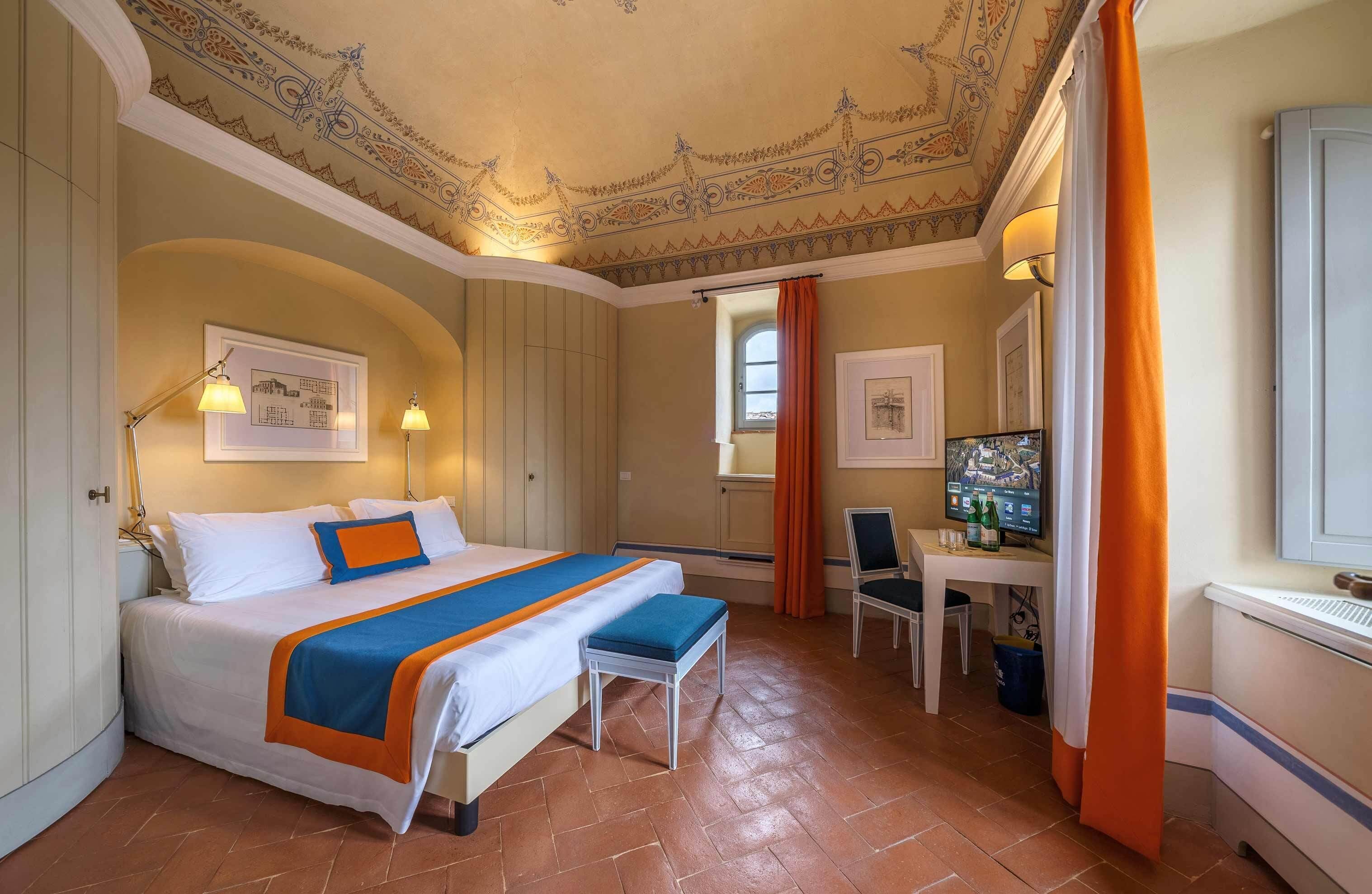 Ein Hotelzimmer mit einem großen Bett, einem Schreibtisch mit Computer, einem Sessel und einem Fenster mit Vorhängen.