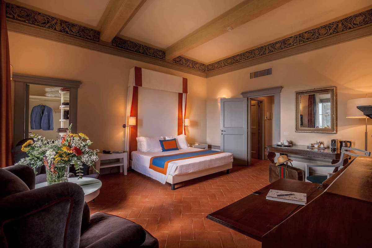 Hotelkamer met een bed, bureau, stoel, tafel, spiegel, garderobe en een bloemenvaas.