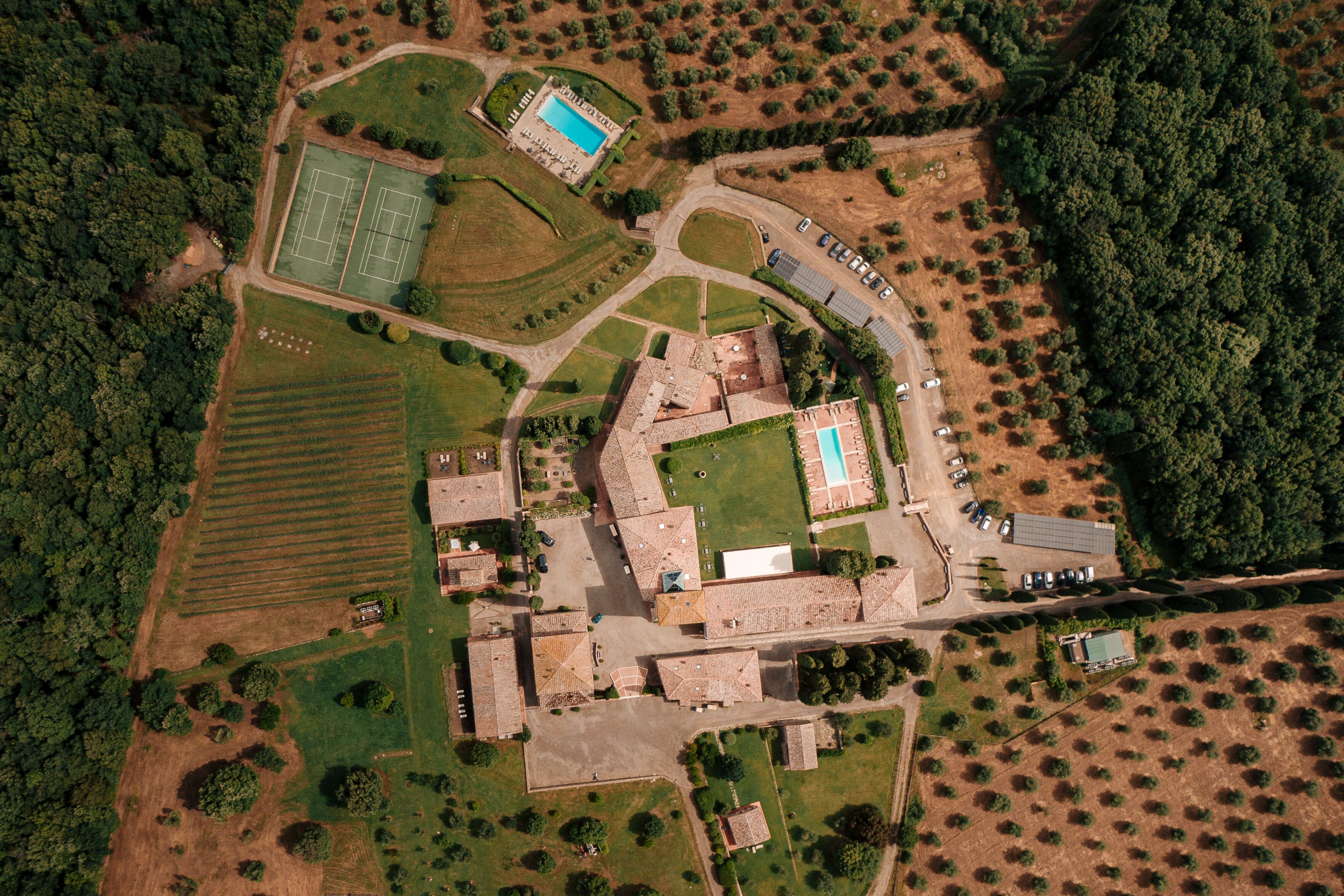 An aerial view of a property with a tennis court, swimming pool, parking area, and surrounding greenery.