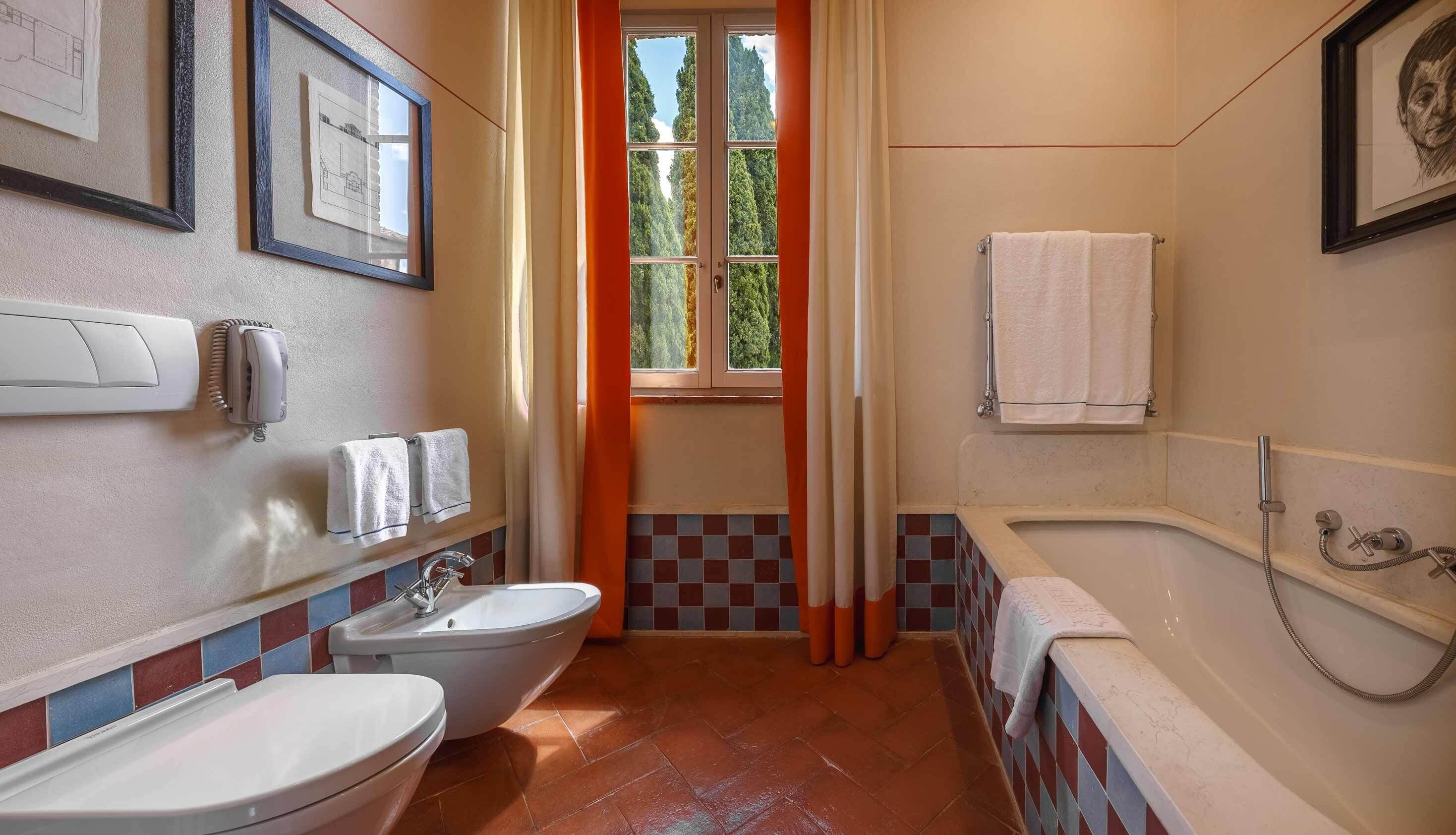 Um banheiro com vaso sanitário, bidê e banheira, uma janela com cortinas e paredes revestidas de azulejos.