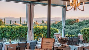 Un área de comedor al aire libre con mesas dispuestas para una comida, un cartel que indica la carta de vinos y vistas del paisaje.