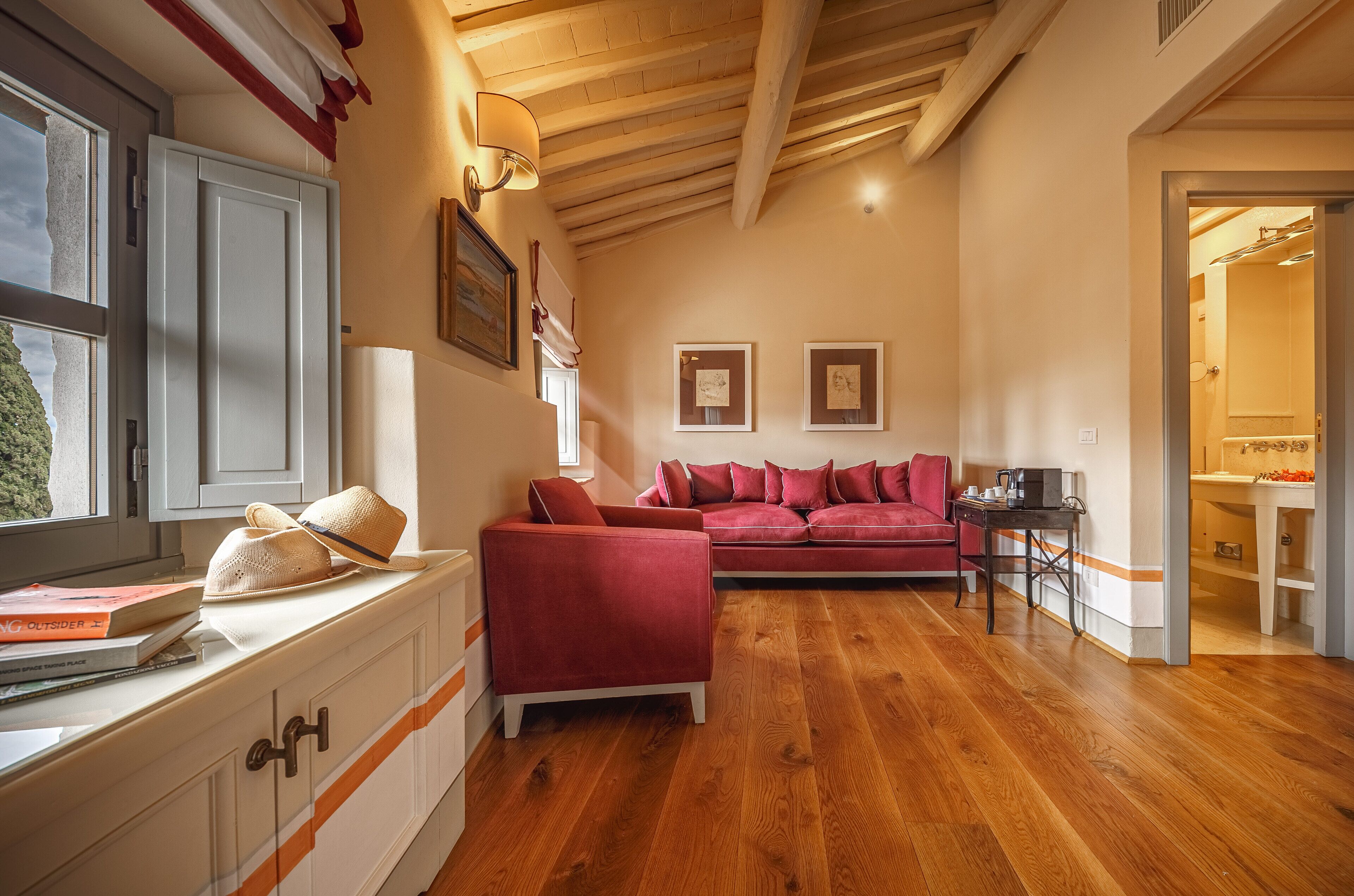 Un salon spacieux avec un plafond en bois, un canapé rouge, une petite table et une fenêtre sur le rebord de laquelle sont posés des livres et un chapeau.