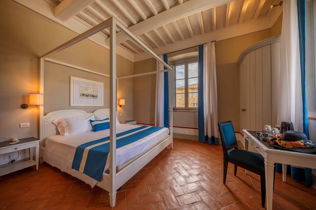 Een hemelbed met een blauw-witte dekbedovertrek, een nachtkastje met een lamp, een stoel en een klein tafeltje met een dienblad.